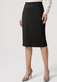 Gonna a matita nera realizzata in tessuto liscio, caratterizzata da cuciture laterali e una silhouette aderente, abbinata a tacchi alti in camoscio grigio chiaro.