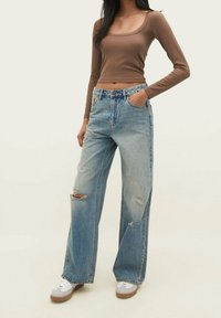 Kobieta ubrana w brązowy długi rękaw crop top, jasnoniebieskie, podarte szerokie jeansy i białe trampki, z jedną ręk�ą w kieszeni.