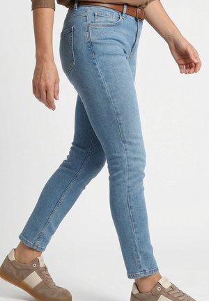 Persoon draagt lichtblauwe skinny jeans, een bruine riem en taupekleurige sneakers en loopt voor een effen achtergrond.