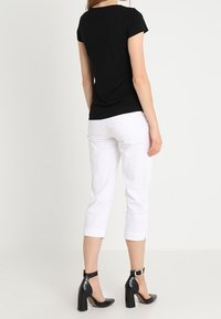 Kobieta ubrana w czarną bluzkę z krótkim rękawem, białe spodnie w kostkę i czarne szpilki na kostkę, stojąca na białym tle.