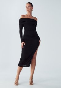 Melna kleita ar atsegtiem pleciem, garām piedurknēm, rušļotu faktūru un augstu šķēlumu līdz augšstilbam. Kopā valkātas ar zelta krāsas siksniņām apavotām papēžiem. Minimālistisks fons.