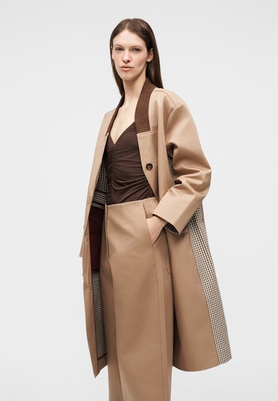 Γυναίκα που φοράει μακρύ μπεζ παλτό με καρό φόδρα, καφέ ριγέ τοπ με ριχτά σχέδια και ταιριαστά μπεζ ψηλόμεσα παντελόνια, με το χέρι στην τσέπη.