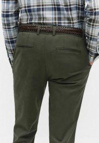 Man som bär olivgröna byxor med bakfickor och ett flätat brunt läderbälte, tillsammans med en rutig skjorta i blått och brunt.