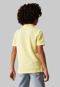 Polo giallo con maniche corte e colletto, caratterizzato da spacco laterale all'orlo. Indossato con jeans in denim blu chiaro.