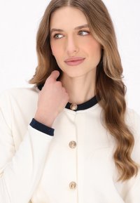 White garment with a navy collar and cuffs, featuring decorative gold buttons. Smooth texture, classic design with a fitted style.