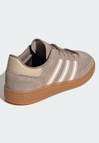 Baskets en cuir suédé marron avec un design à trois bandes blanches, un talon en cuir beige et une semelle en caoutchouc gomme texturée. Comprend un col rembourré.
