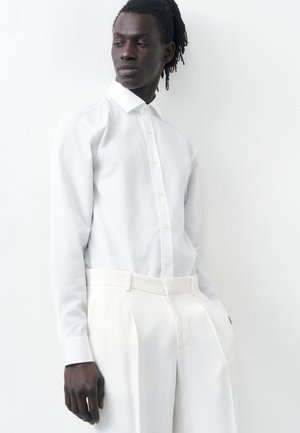 Moški v beli srajci z gumbi in belih hlačah z visokim pasom, stoji z rokami v žepih pred belim ozadjem.