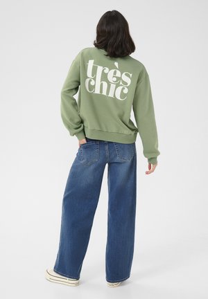 Person med skulderlangt mørkt hår iført grøn sweatshirt med teksten "très chic" på ryggen, vide blå jeans og hvide sneakers.