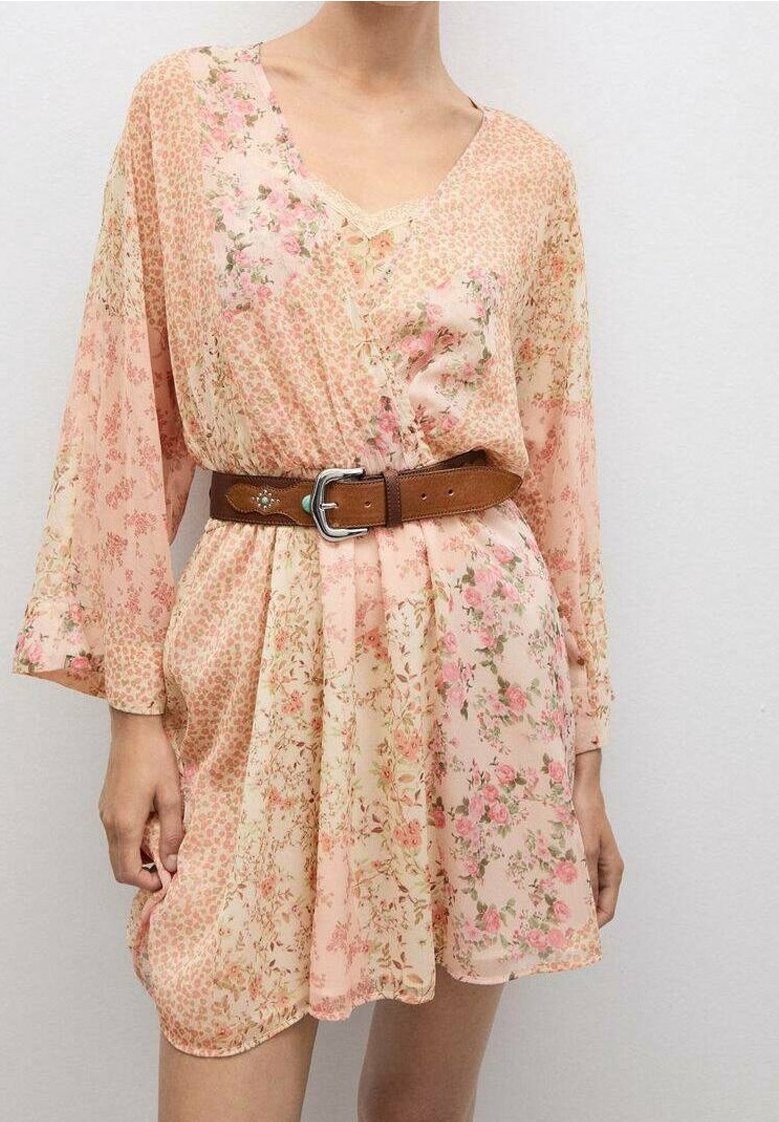 Femme portant une robe rose clair à motif floral avec des manches longues et une ceinture marron à la taille sur un fond uni.
