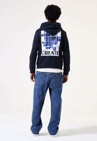 Sudadera navy con un diseño gráfico en azul y blanco, que presenta la palabra "CREATE". Combinada con unos vaqueros de denim azules de corte holgado.