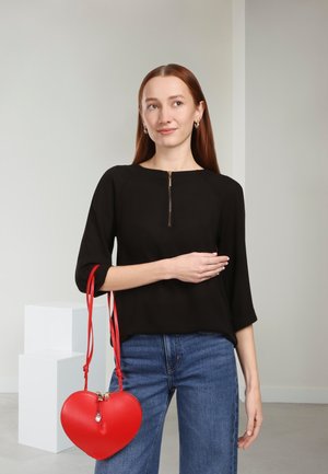 Zwart topje met een rits aan de voorkant, gecombineerd met high-waisted blauwe jeans. Een rode handtas in de vorm van een hart met een rits hangt aan een schouderriem.