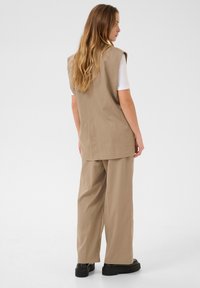 Beige oversized vest with no sleeves paired with loose-fitting beige trousers. Smooth texture, minimalistic design, and black platform shoes.