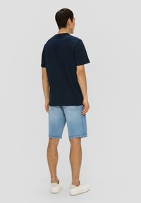 Marineblauw katoenen t-shirt met korte mouwen, gecombineerd met lichtblauwe denim shorts. Model in witte sneakers, gezien vanaf de achterkant.