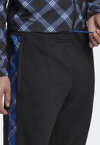 Pantaloni sportivi neri con una striscia laterale blu caratterizzata da un motivo a quadri. Il tessuto sembra morbido; la vita include un cordoncino per la regolazione.