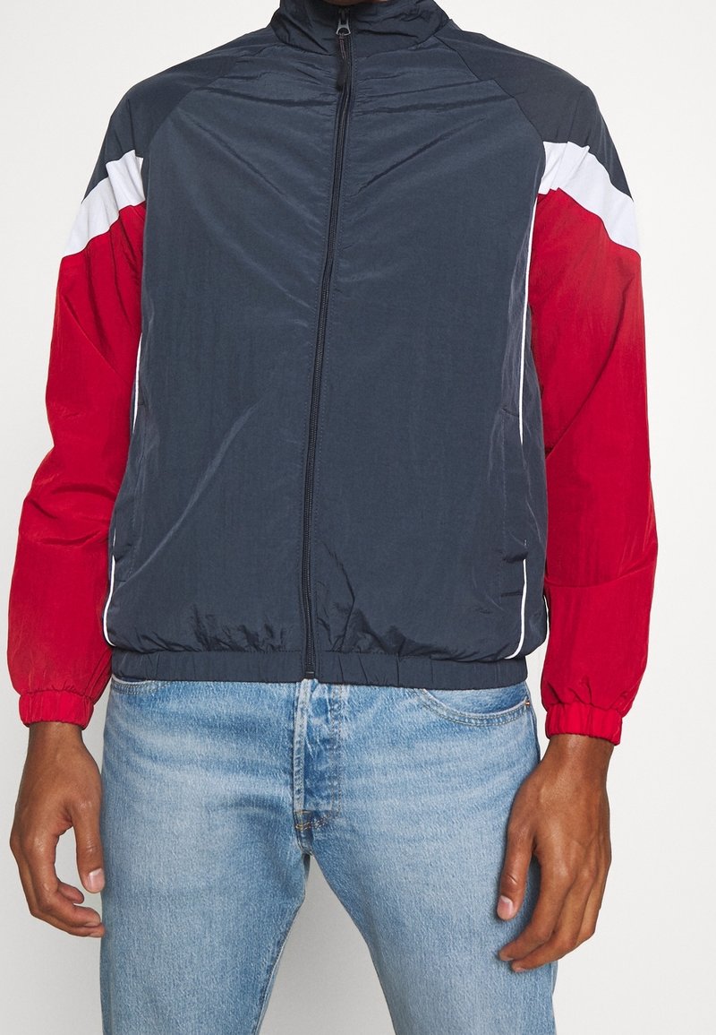 Merin ja punainen tuulitakki, jossa on valkoisia yksityiskohtia; varustettu vetoketjukiinnityksellä, seisomakauluksella ja joustavuusrannekeilla. Kevyt, sileä tekstuuri.