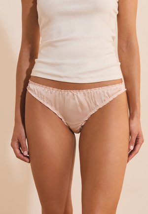 Frau trägt hellrosa Satin-Bikini-Unterwäsche und ein weißes ärmelloses Top, stehend vor einem beigen Hintergrund.