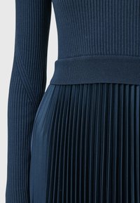 Laivastonsininen mekko, jossa on ribbetty yläosa ja laskostettu hame. Materiaali on sileä ja teksturoitu yläosa. Muotoon ommeltu, jossa on vyötärökaista.