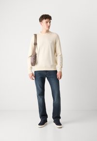 Light beige sweater with ribbed hem, paired with dark blue jeans. Model also carries a small gray crossbody bag and wears navy sneakers.