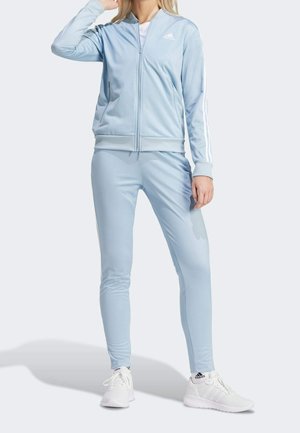Kvinde iført en lyseblå Adidas træningsdragt med hvide striber og hvide Adidas sneakers, der står foran en ensfarvet baggrund.