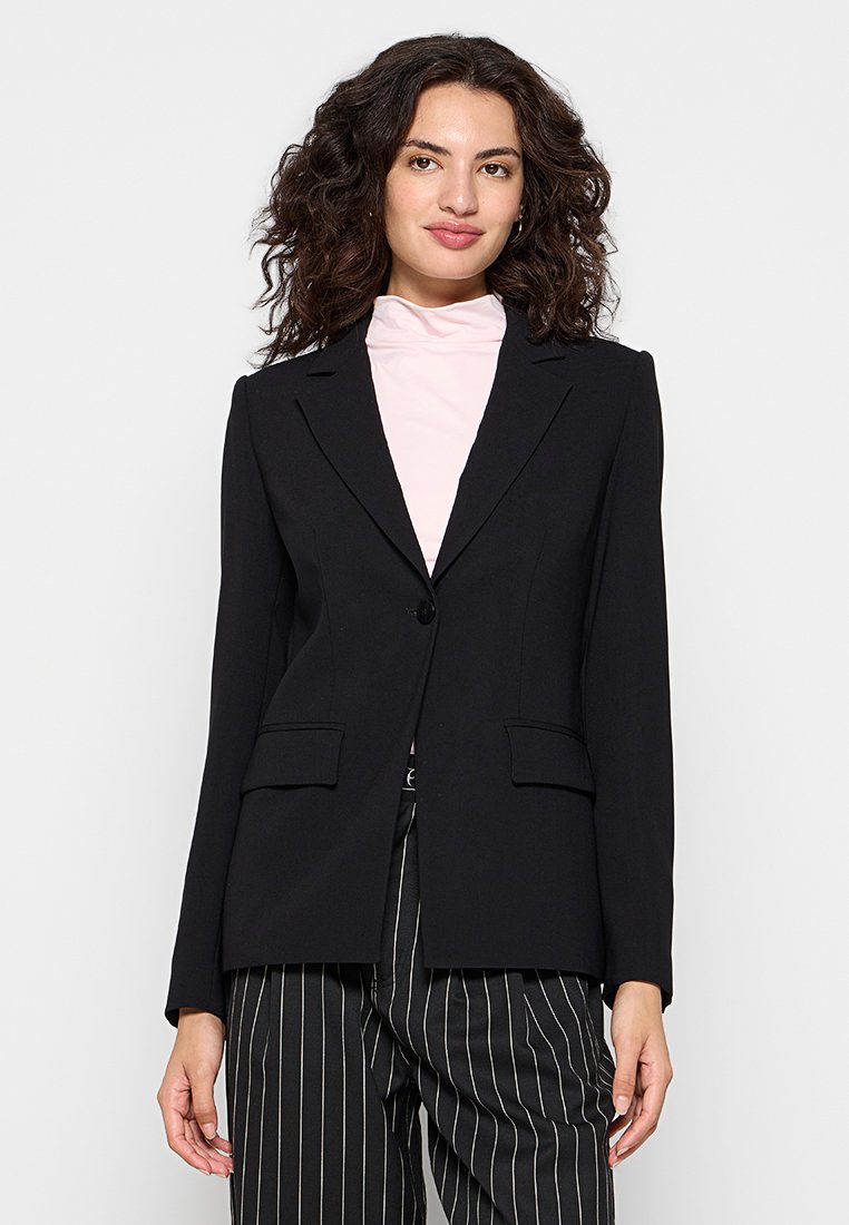 Blazer noir ajusté avec des manches longues, revers à cran, fermeture à un bouton et deux poches avant. Porté par-dessus une chemise rose clair.