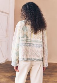 Mehrfarbige gemusterte Jacke mit gesteppter Textur, mit geometrischen Designs in Grün, Blau, Pink und Gelb auf hellem Untergrund.