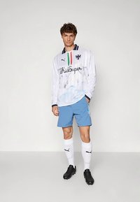 Junger Mann steht und trägt ein weißes langärmliges Fußballtrikot, blaue Shorts, weiße kniehohe Socken und schwarze Fußballschuhe vor einem einfarbigen Hintergrund.