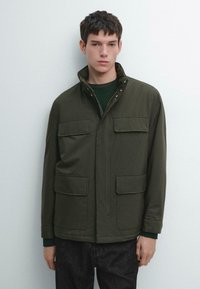 Giacca verde scuro con zip e collo, due tasche sul petto e tessuto morbido. Indossata sopra un maglione verde e abbinata a jeans.