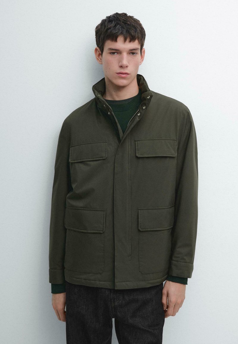 Giacca verde scuro con zip e collo, due tasche sul petto e tessuto morbido. Indossata sopra un maglione verde e abbinata a jeans.