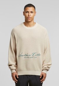 Beige striktrøje med afslappet pasform, ribbede halsudskæring og manchetter. Har broderet tekst "Another Cotton Established 2023" i teal.
