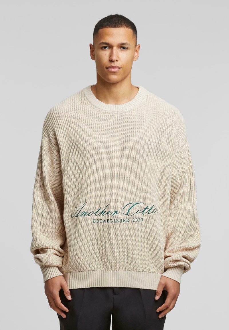 Beige striktrøje med afslappet pasform, ribbede halsudskæring og manchetter. Har broderet tekst "Another Cotton Established 2023" i teal.