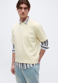 Νεαρός άνδρας που φορά μπλούζα κοντομάνικη ανοιχτού κίτρινου χρώματος πάνω από ριγωτό πουκάμισο με μακριά μανίκια και τζιν ανοιχτού μπλε χρώματος, στέκεται μπροστά σε λευκό φόντο.