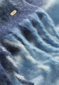Blå ombre tørklæde lavet af blødt, fuzzy materiale. Har frynsede kanter med varierende nuancer af blå og et lille metallic logo tag.