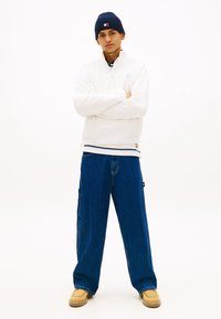 Λευκό πουλοβέρ με φερμουάρ στο ένα τέταρτο, φορεμένο πάνω από μπλε μακρυμάνικο με ρίγες, συνδυασμένο με φαρδιά μπλε τζιν παντελόνια και ταμπά μοκασίνια.