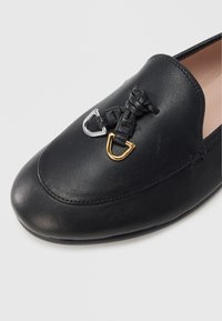 Scarpa nera in pelle con punta arrotondata, caratterizzata da dettagli intrecciati e due accessori metallici in argento e oro. Tessitura liscia.