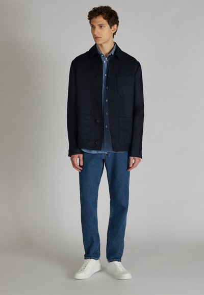 Veste marine avec un col, une poche avant et une fermeture à boutons, superposée à une chemise en denim. Portée avec un jeans bleu et des baskets blanches.
