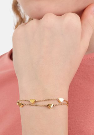 Bracciale dorato con due catene, caratterizzato da cinque piccoli charm a forma di cuore lucidi. Il bracciale è indossato su un polso su uno sfondo rosa.