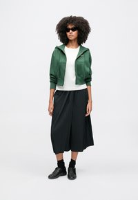 Vrouw met krullend haar die een zonnebril draagt, een groene capuchontrui met ritssluiting, wit shirt, zwarte geplooide broek met wijde pijpen, zwarte sokken en zwarte schoenen.