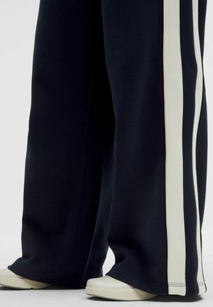 Széles szárú fekete nadrág két függőleges fehér csíkkal az oldalán, fehér lapos cipővel párosítva, világos háttéren.