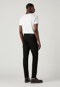 Bărbat în picioare, de profil, purtând un tricou alb, blugi negri, șosete albe și pantofi eleganți maro, pe un fundal simplu.