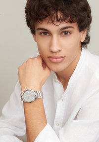 Montre-bracelet analogique argentée avec un cadran bleu clair, un bracelet en acier inoxydable et des index dorés. Le modèle porte une chemise en lin blanche.