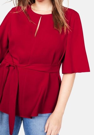 Blouse rouge avec une découpe en forme de goutte d'eau au niveau du col, manches courtes évasées et taille nouée pour une silhouette cintrée. Tissu lisse avec une coupe décontractée.