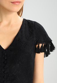 Vestido negro con escote en V y mangas cortas, que presenta un tejido texturizado, patrones ornamentales y acentos de borlas negras en las mangas.