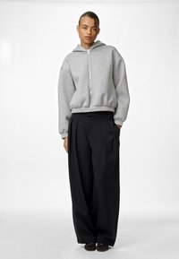 Siva dukserica na patent zatvarač izrađena od mekog materijala, sa kapuljačom i spuštenim ramenima, u kombinaciji sa širokim crnim hlačama. Odjeća ima opušteni kroj.