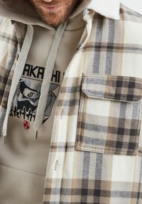 Camicia in flanella a scacchi nei toni crema, marrone e grigio con colletto a contrasto; presenta due tasche sul petto. Sotto c'è un hoodie beige con stampa grafica.