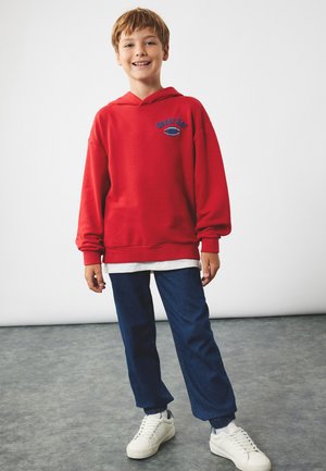 Lächelnder Junge mit kurzen Haaren, der einen roten Kapuzenpullover mit dem Text "Toller Tag", blaue Hosen und weiße Turnschuhe trägt und auf einem grauen Boden steht.