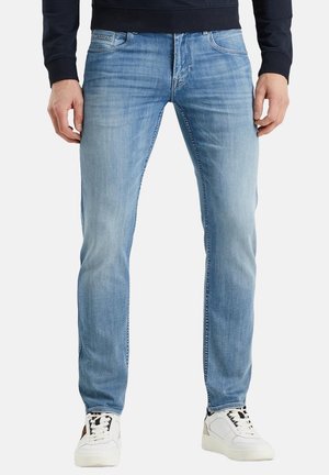 Mann trägt hellblaue Slim-Fit-Jeans und weiße Sneaker, Hände locker an den Seiten, vor schlichter Hintergrund.