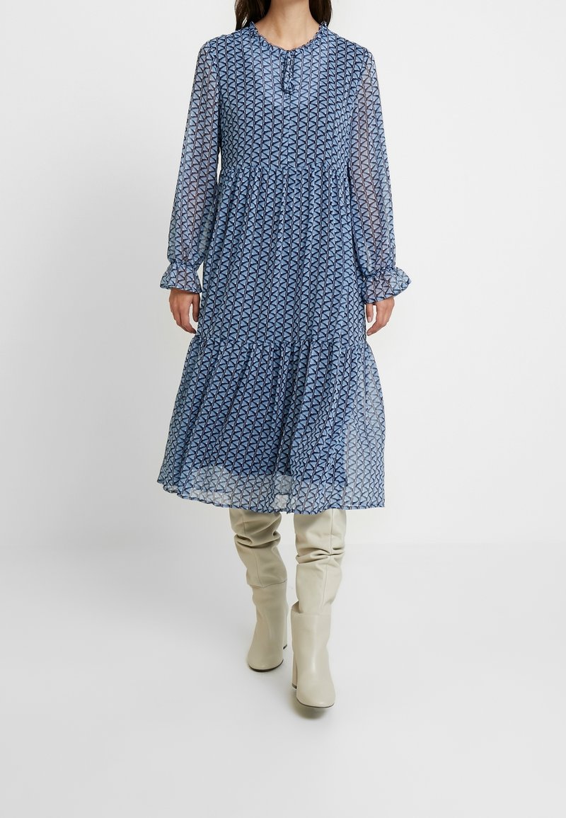 Robe midi à motifs bleus avec de longues manches transparentes, un col rond et une jupe à volants. Portée avec des bottes crème à hauteur de genou décontractées.