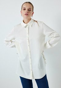 Camisa blanca de manga larga hecha de una tela suave, con cierre de botones en el frente, cuello clásico y puños fruncidos para un detalle adicional.