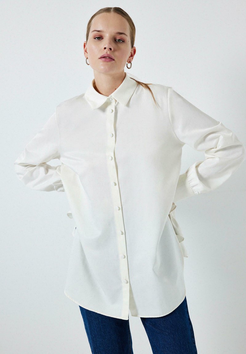 White long-sleeve shirt made of a smooth fabric, featuring a button-down front, classic collar, and ruffled cuffs for added detail.