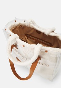 Transparent tote-väska med creamfärgad faux shearling-foder, bruna läderhandtag och en inre dragkedjeficka. Broderad logotyp på utsidan.